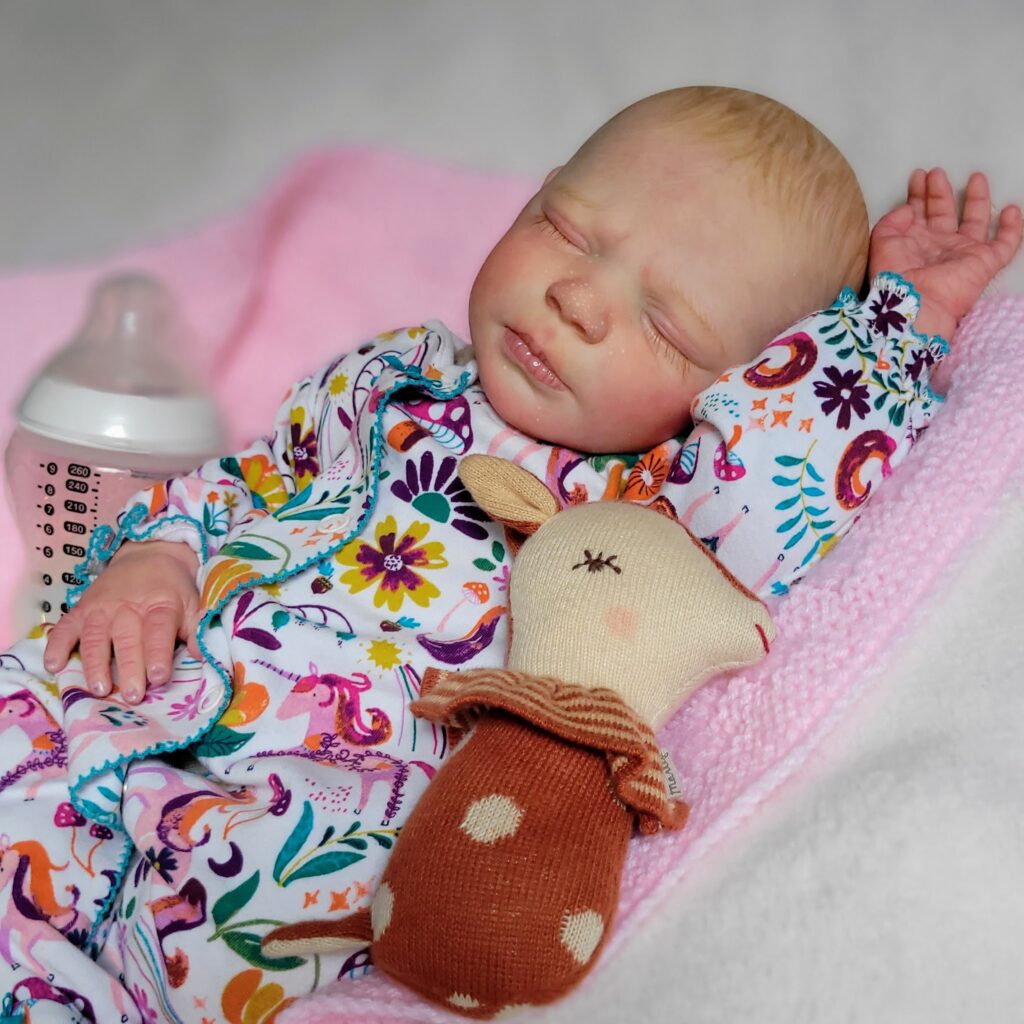 Realistic reborn doll sleeping in floral sleepsuit with soft toy and bottle on pink blanket