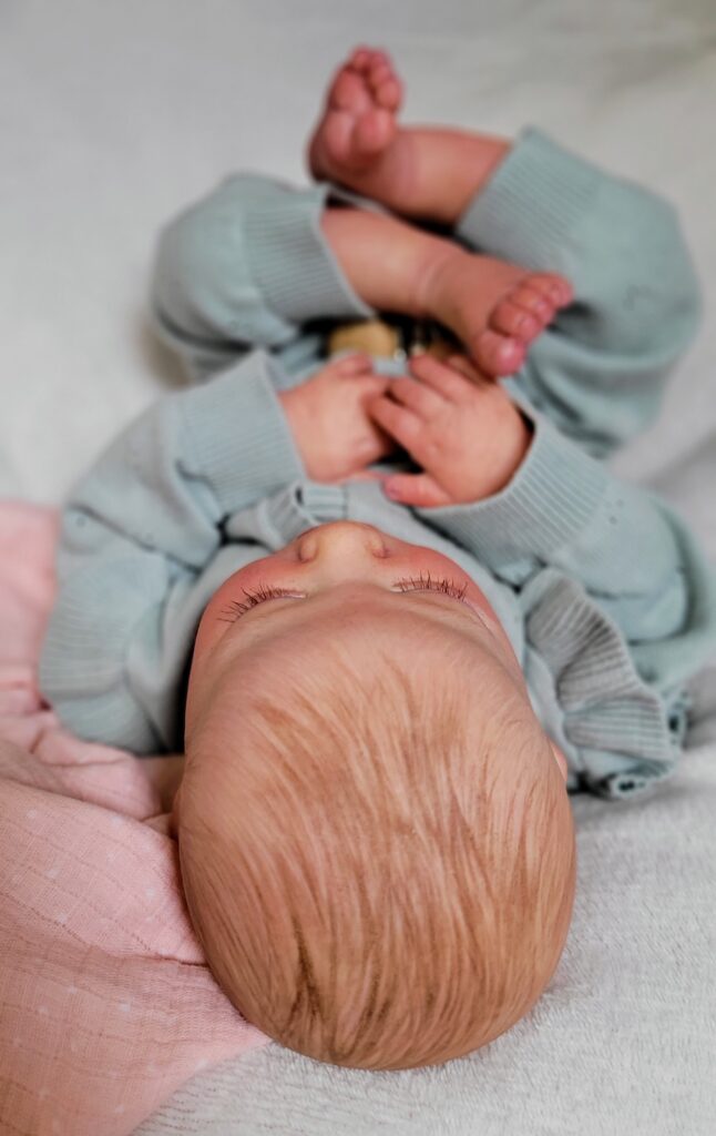 Reborn doll in soft green knitted outfit holding feet, lying on a pink muslin blanket — top-down view