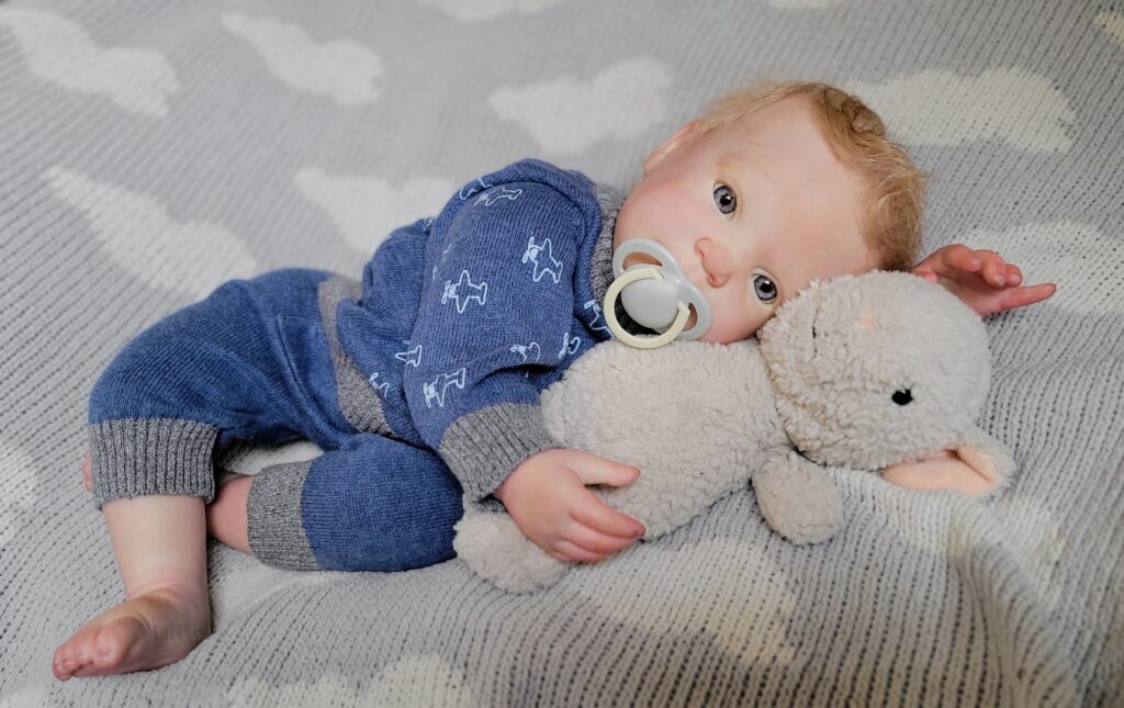 reborn baby doll with blue eyes and dummy cuddling a plush lamb on a grey cloud blanket